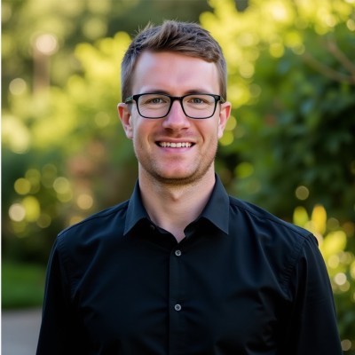 Portrait of Jared Verbeke smiling, wearing glasses and a black shirt, with a blurred green outdoor background.